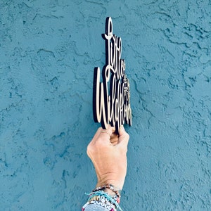 May include: Wooden sign with the words "Little Wildflower" in a cursive font. The sign is held against a textured blue wall. The sign is made of light-colored wood with a dark border.
