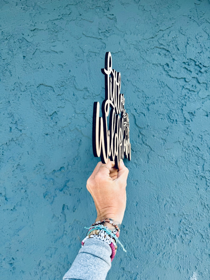 May include: Wooden sign with the words "Little Wildflower" in a cursive font. The sign is held against a textured blue wall. The sign is made of light-colored wood with a dark border.