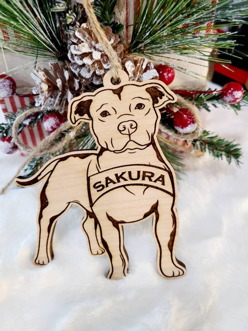 K&ouml;nnte beinhalten: Ein h&ouml;lzernes Hundeornament mit dem Namen "SAKURA" auf einem Banner eingraviert. Das Ornament ist hellbraun mit dunkleren braunen ge&auml;tzten Details und h&auml;ngt vor einer Weihnachtsdekoration mit Tannenzapfen und roten Beeren.