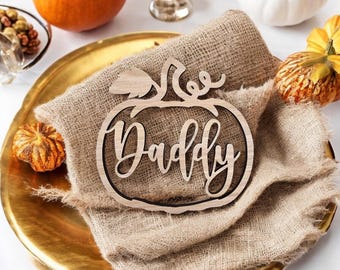 May include: Wooden pumpkin-shaped place card with the word "Daddy" written in a decorative font. The place card is on a hessian napkin.