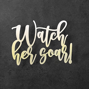 May include: A decorative sign with the words "Watch her soar!" in a cursive font. The text is a gradient of gold and white, set against a dark gray, textured background.