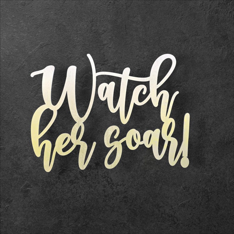 May include: A decorative sign with the words "Watch her soar!" in a cursive font. The text is a gradient of gold and white, set against a dark gray, textured background.