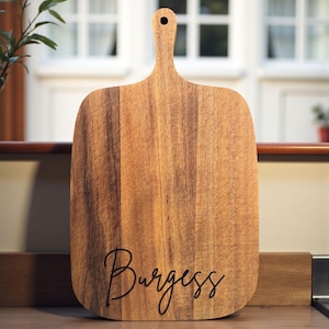 A wooden cutting board with the word "Burges" written on it, placed on a wooden surface.