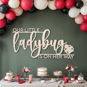 May include: A wooden sign that reads "OUR LITTLE ladybug IS ON HER WAY" is displayed on a green wall. The sign is decorated with a ladybug graphic. Balloons in red, white, and black are arranged above the sign. Cakes and flowers are on a table.