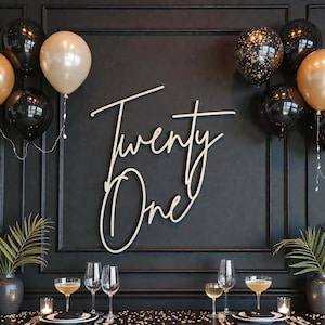 Puede incluir: Una pared negra con un letrero de madera que dice "Twenty One" en escritura cursiva. La pared est&aacute; decorada con globos negros y dorados, y una mesa est&aacute; preparada para una celebraci&oacute;n con copas y velas.