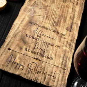 May include: A wooden serving board with engraved text. The text reads "Never underestimate the difference you have made and the lives you have touched. Happy Retirement." The board is light brown with a natural wood grain.