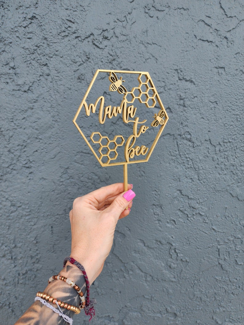 May include: A gold wooden hexagonal sign with the text "Mama to bee" and two bee illustrations. The sign is held in a hand.