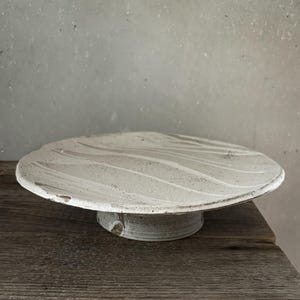 Könnte beinhalten: Handgefertigte, cremefarbene Keramik-Kuchenplatte mit strukturiertem, wellenförmigem Design auf der Platte. Der Ständer hat einen kreisförmigen Sockel und einen leicht gewellten Rand. Geeignet zum Präsentieren von Backwaren.