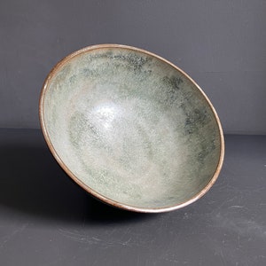 May include: A large, round, green ceramic bowl with a brown rim. The bowl is slightly tilted and has a speckled, textured surface.