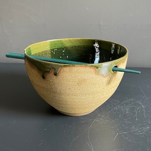 May include: A green and brown ceramic bowl with a hole in the side for chopsticks. The bowl is sitting on a dark gray surface.