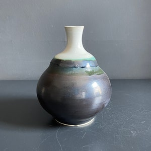 May include: A small, round, ceramic vase with a narrow neck. The vase has a white glaze on the top and a dark brown glaze on the bottom, with a band of green and blue glaze in between.