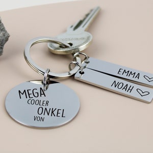 Könnte beinhalten: Silberner Schlüsselanhänger mit einem runden Anhänger, auf dem "MEGA COOLER ONKEL VON" steht, und zwei rechteckigen Anhängern, auf denen "EMMA ♥" und "NOAH ♥" steht.