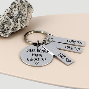 Könnte beinhalten: Silberner Schlüsselanhänger mit einem runden Anhänger, auf dem "Diese Bonus Mama gehört zu" mit einem Herzsymbol steht. Es sind drei rechteckige Anhänger an dem Schlüsselanhänger befestigt, auf denen "Cody", "Luke" und "Lias" mit Herzsymbolen stehen.