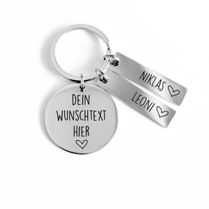 Könnte beinhalten: Silberner Schlüsselanhänger mit einem runden Anhänger, auf dem "Dein Wunschtext Hier" steht, und zwei kleineren Anhängern, auf denen "Niklas" und "Leoni" mit einem Herzsymbol stehen.