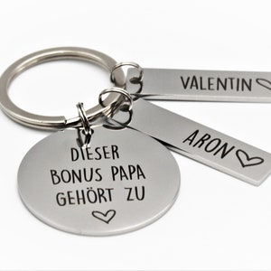 Könnte beinhalten: Silberner Schlüsselanhänger mit einem runden Anhänger, auf dem "Dieser Bonus Papa gehört zu" mit einem Herzsymbol steht. Zwei kleinere Anhänger sind am Schlüsselanhänger befestigt, einer mit der Aufschrift "Valentin" mit einem Herzsymbol und der andere mit der Aufschrift "Aron" mit einem Herzsymbol.