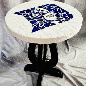 Op de afbeelding: Een ronde witte tafel met een blauw mozaïekontwerp van een kattengezicht. De tafel heeft een zwarte houten basis met een uniek ontwerp.