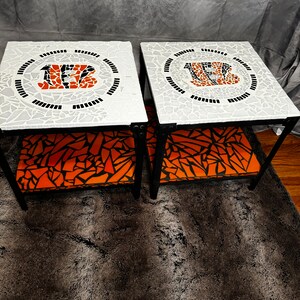 Op de afbeelding: Twee zwarte metalen bijzettafels met oranje en zwarte mozaïektegels met het logo van de Cincinnati Bengals. De tafels hebben oranje en zwarte gepatroneerde stoffen planken.