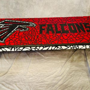 Op de afbeelding: Een mozaïek salontafel met een rood, wit en zwart ontwerp. Het tafelblad heeft een zwart-witte afbeelding van een valk met het woord "FALCONS" in zwarte letters. De tafel heeft zwarte poten.