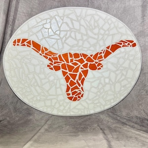 Op de afbeelding: Een witte ronde mozaïektegel met een oranje longhorn stierenkopontwerp.