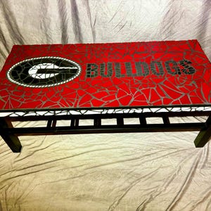 Op de afbeelding: Een mozaïek salontafel met een rood en zwart logo van de University of Georgia Bulldogs. De tafel heeft een zwart metalen frame met een plank.