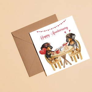 May include: A greeting card with a happy anniversary message. Two dachshund dogs are sitting at a table with a cup of coffee and a heart-shaped balloon. The card has a string of red hearts across the top.