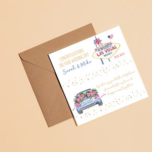 May include: A white wedding card with gold glitter and a watercolor illustration of a car decorated with flowers. The card says "Congratulations on your wedding day Sarah & Mike" and "Wishing you both a lifetime of everlasting love & happiness together". The card also features a sign that says "Welcome to Fabulous Las Vegas Nevada" with a red heart and a small American flag.