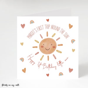 Op de afbeelding: Een witte kaart met een aquarel-illustratie van een lachende zon in rood, oranje en gele tinten. De tekst "MARGOT'S FIRST TRIP AROUND THE SUN" staat in een cirkel rond de zon. De tekst "Happy 1st Birthday little one" staat onder de zon. De kaart is versierd met kleine rode hartjes en regenboog-illustraties.