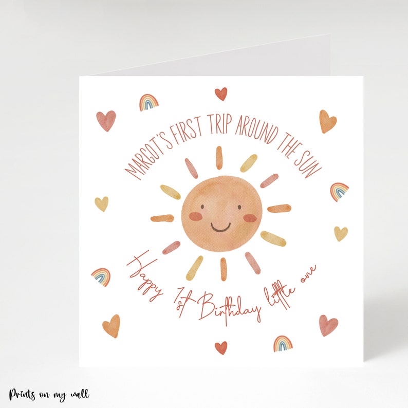 Op de afbeelding: Een witte kaart met een aquarel-illustratie van een lachende zon in rood, oranje en gele tinten. De tekst "MARGOT'S FIRST TRIP AROUND THE SUN" staat in een cirkel rond de zon. De tekst "Happy 1st Birthday little one" staat onder de zon. De kaart is versierd met kleine rode hartjes en regenboog-illustraties.