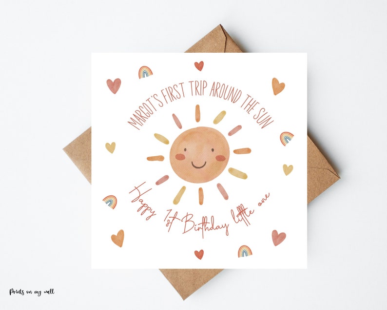 Op de afbeelding: Een witte kaart met een zonillustratie en de tekst "MARGOT'S FIRST TRIP AROUND THE SUN Happy 1st Birthday little one". De zon is oranje en geel met een glimlachend gezicht. Rond de zon zijn kleine rode hartjes en regenboogillustraties te zien.