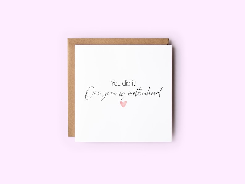 May include: A square greeting card with a brown envelope. The white card has the text "You did it! One year of motherhood" in black script, with a small pink heart below the text. The background is a light pink.