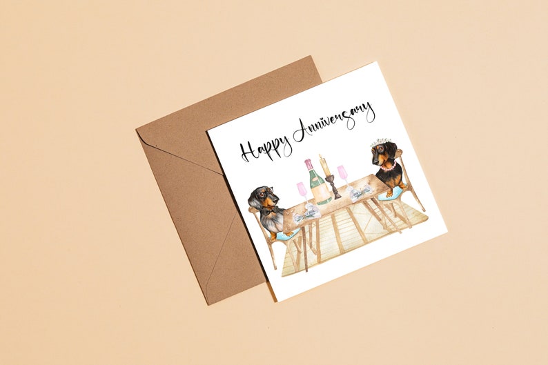 May include: A greeting card with a watercolor illustration of two dachshunds at a table set for a celebration. The card reads "Happy Anniversary" in elegant script. A brown envelope is next to the card.