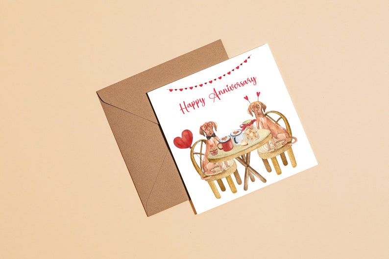 K&ouml;nnte beinhalten: Eine wei&szlig;e Karte mit einer roten Herzgirlande und dem Text "Happy Anniversary". Zwei braune Hunde sitzen an einem Tisch mit einer roten Tasse und einem Teller mit Leckerlis. Die Hunde tragen rote Fliegen.