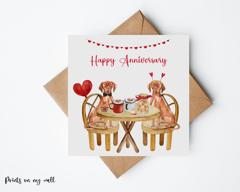 K&ouml;nnte beinhalten: Eine Gru&szlig;karte mit einer Aquarell-Illustration von zwei Hunden, die Herz-H&uuml;te tragen und an einem Tisch mit Kaffeetassen und Keksen sitzen. Die Karte lautet "Happy Anniversary".