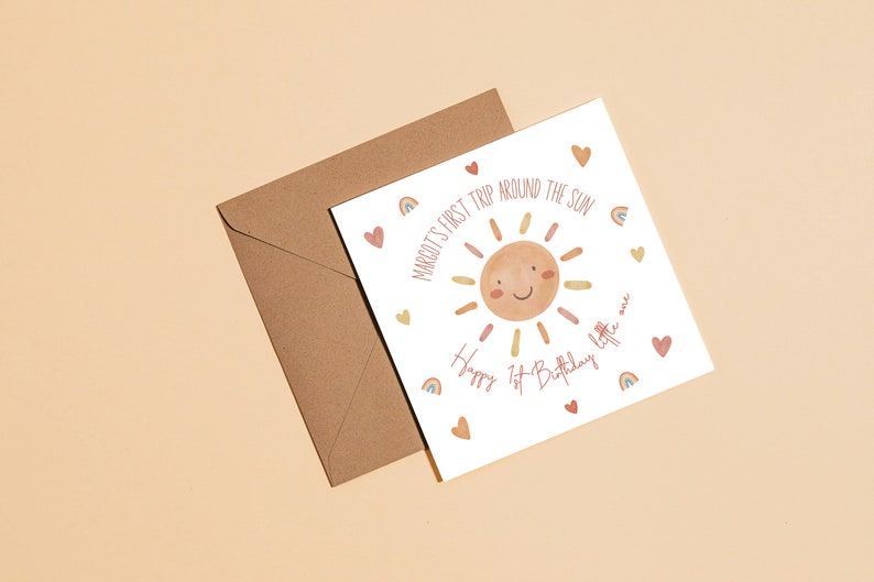 Op de afbeelding: Een witte kaart met een zonillustratie en de tekst "Margot's First Trip Around The Sun Happy 1st Birthday Little One" met regenboog- en hartachtige accenten.