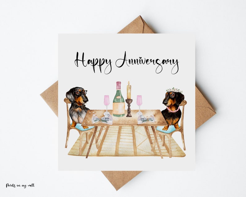 May include: A square greeting card with a watercolor illustration of two dachshunds at a table set for a romantic dinner. The card reads "Happy Anniversary" in elegant script. A brown envelope is behind the card.