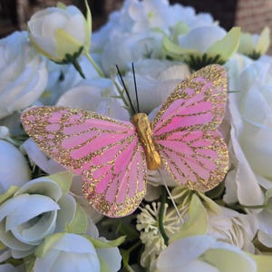 May include: A pink butterfly decoration with gold glitter accents on its wings. The butterfly has a gold body and black antennae. It is set against a backdrop of white and light green flowers.