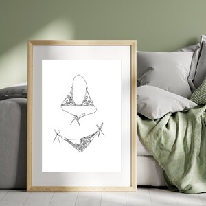 May include: A black and white line drawing of a bikini with floral details. The drawing is framed and displayed on a wall.