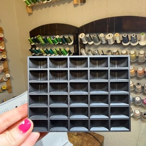 May include: A dark gray, 3D-printed organizer with 30 square compartments. The organizer is held in front of a background with spools of thread in various colors. The organizer is designed to hold small items.