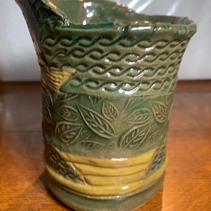 May include: A green ceramic pitcher with a textured surface. The pitcher has a wide mouth and a handle. The pitcher is decorated with a pattern of leaves and a band of yellow glaze.