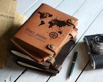 May include: A brown leather travel journal with a world map and compass design. The journal has the text "Your name - Travel Planner" on the front cover.