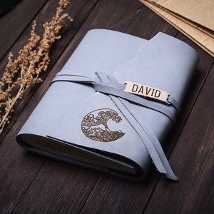 May include: A light blue leather journal with a tree design and the name "DAVID" engraved on a leather tag. The journal is tied with a leather strap.