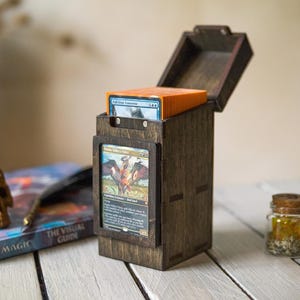 May include: Wooden card storage box with an open lid, holding a stack of orange-edged cards. A Magic: The Gathering card is visible in the front. Small glass jars with cork lids and dice are in the background. A skull and book are also present.