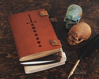 May include: A brown leather journal with a sword and dice design, stacked on top of two other journals. The journal is on a wooden surface with two skulls and a quill pen.