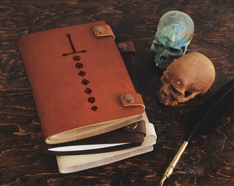 May include: Leather-bound journals with a sword and dice design, featuring a brown, dark brown, and cream color scheme. Two decorative skulls and a quill pen are also present, set against a dark wood background.