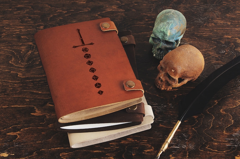 Op de afbeelding: Leren notitieboeken in bruin, donkerbruin en cr&egrave;me, versierd met een zwaard- en dobbelsteenontwerp. Twee decoratieve schedels en een ganzenveer zijn ook aanwezig, tegen een donkere houten achtergrond.