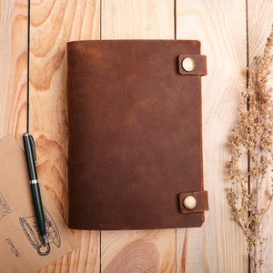 K&ouml;nnte beinhalten: Ein braunes Lederjournal mit zwei Messingverschl&uuml;ssen und einem Stift auf einer Holzoberfl&auml;che.