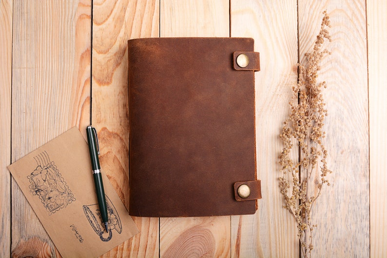 K&ouml;nnte beinhalten: Ein braunes Lederjournal mit zwei Messingverschl&uuml;ssen und einem Stift auf einer Holzoberfl&auml;che.