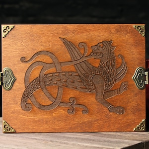 May include: A wooden box with a hinged lid and brass corner accents. The lid features an intricate carved design of a winged creature with a long tail.