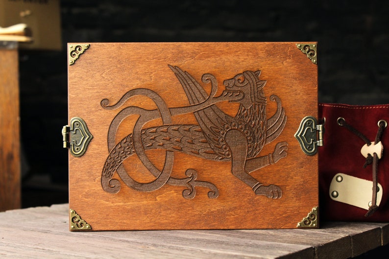 May include: A wooden box with a hinged lid and brass corner accents. The lid features an intricate carved design of a winged creature with a long tail.