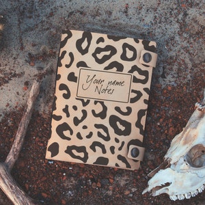 Puede incluir: Un cuaderno marrón con una cubierta de estampado de leopardo. El cuaderno tiene una etiqueta que dice "Your name Notes". El cuaderno está sobre una superficie marrón y gris con un cráneo de ciervo y cuernos.